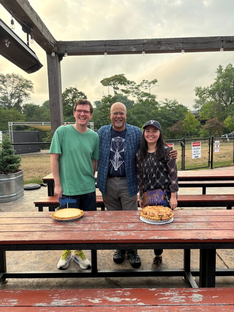 A photo of a professor and two trainees standing behind a table of two pies at a going away celebration.