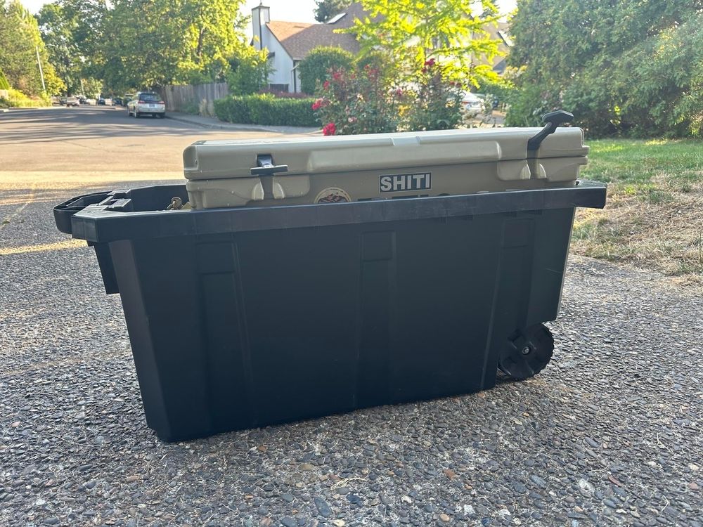 A black plastic storage bin with wheels is placed on a driveway, holding a tan cooler labeled "SHITI."
