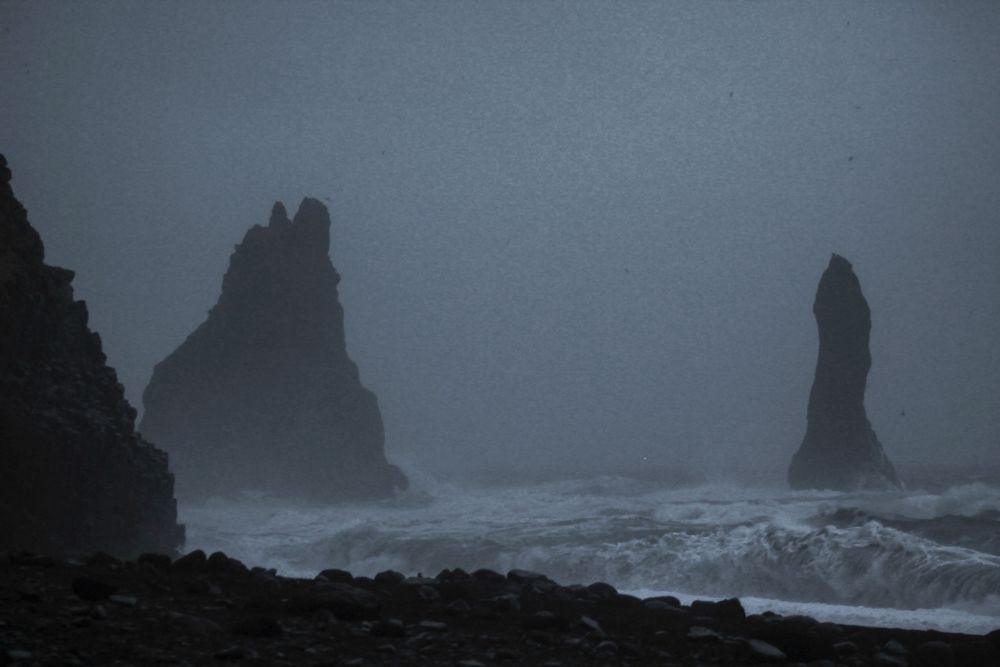 Looking out to see across a black beach. Two outcrops of rock protruding over the waves in a storm.