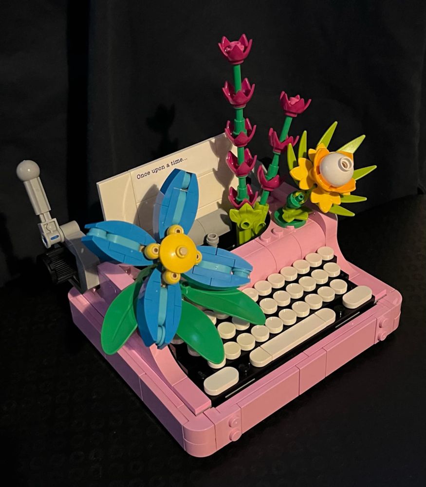A small pink Lego typewriter with white keys. The piece of “paper” in the typewriter reads “Once upon a time.” Flowers grow out of the typewriter’s carriage: a blue, four-petal flower on the left, a large yellow flower on the right, and two spikes of dark pink flowers behind the yellow flower.