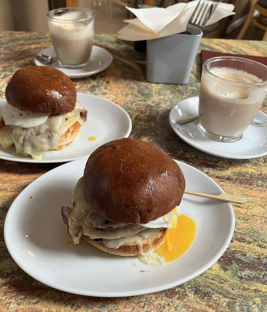 A brioche bun containing sausage patties, bacon, egg, and Swiss cheese, sitting on a white plate. The egg’s yolk has broken and run down onto the plate. Behind it are two glass mugs containing lattes and another breakfast bun (the same contents as the first)