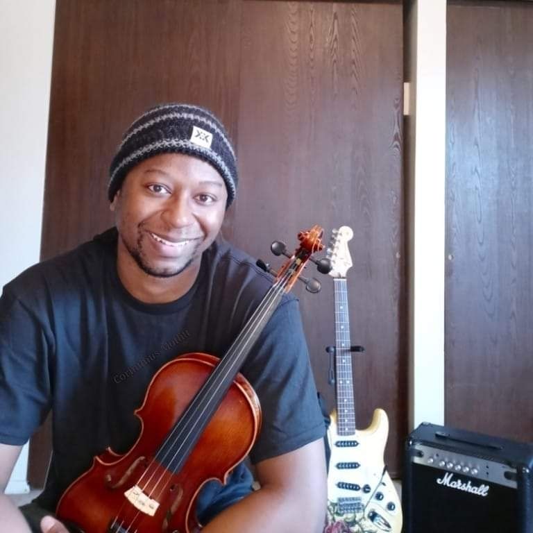 Picture of a smiling handsome man holding a #violin with a #guitar and #amplifier in the background.