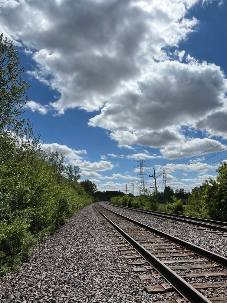 Railroad tracks curving into unit distance among greenery and power lines, bright blue sky with fluffy white clouds overhead