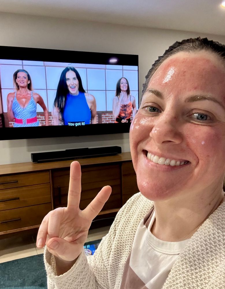 Lady with very red, shiny face making a peace sign in front of tv screen showing movie The Substance 