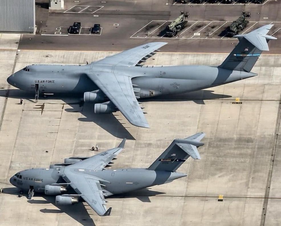 This image shows two large U.S. Air Force cargo aircraft parked on an airbase tarmac. The aircraft at the top is a Lockheed C-5 Galaxy, and the one at the bottom is a Boeing C-17 Globemaster III. Here’s a quick comparison and description:

Top Aircraft: C-5 Galaxy
	•	One of the largest military aircraft in the world.
	•	Distinctive features:
	•	High T-tail.
	•	Four turbofan engines mounted on pylons beneath a high-mounted wing.
	•	Long fuselage with a pronounced hump at the front.
	•	Nose section can lift up for front loading.
	•	Used for strategic airlift missions.

Bottom Aircraft: C-17 Globemaster III
	•	A smaller, more modern strategic and tactical airlifter.
	•	Features:
	•	Also has four turbofan engines.
	•	T-tail like the C-5 but with a more compact, streamlined body.
	•	Rear-loading ramp.
	•	Known for its ability to operate from short and unprepared airfields.

Visual Comparison
	•	The C-5 Galaxy is significantly larger in length and wingspan.
	•	The C-17 is more compact and versatile for various airlift operations.
	•	Both aircraft serve critical roles in the U.S. military’s ability to deploy and sustain forces worldwide.

Also visible in the background are some military vehicles and a few parked cars, suggesting this is part of a larger airbase complex.