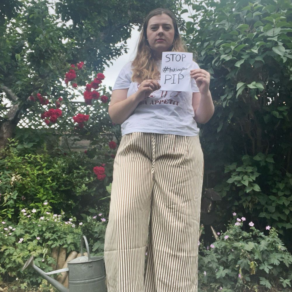 Laura, a white woman with long light brown hair, stands in her garden holding a sign reading ‘Stop taking the PIP’. 
