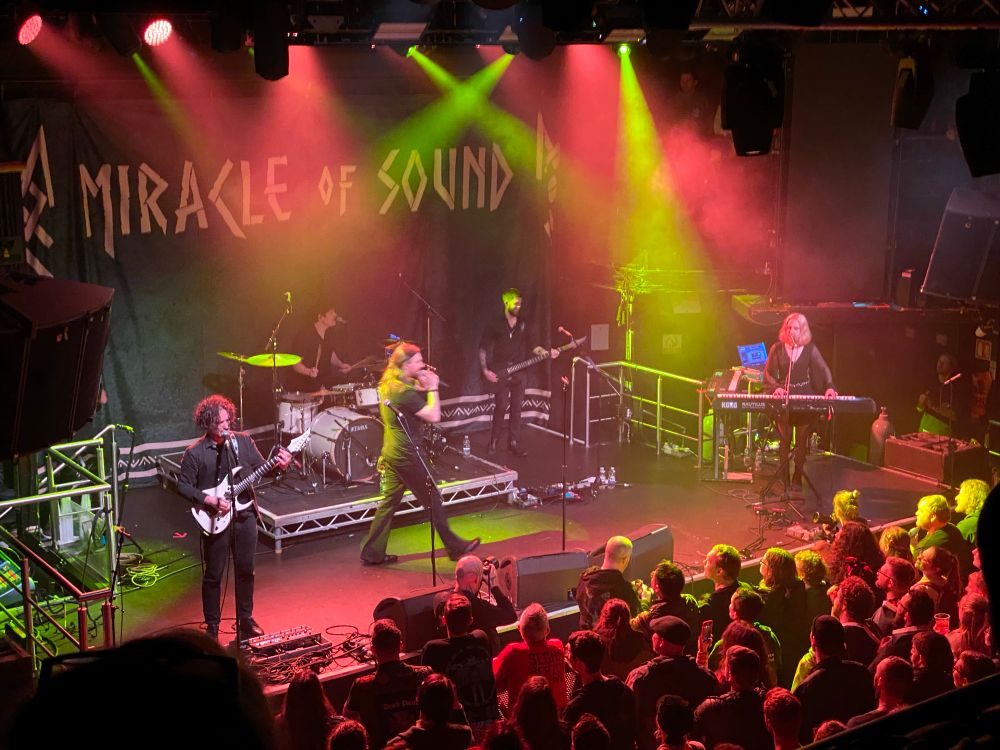 A band play on a stage before an audience of hundreds, all bathed in red and yellow stage lights. A banner at the back of the stage proclaims this band to be Miracle of Sound.

For the uninitiated, Miracle of Sound is the long running solo project of Gavin Dunne, the long haired dude striding across the front of the stage, who writes songs in various styles inspired by video games, shows, movies and books. He liked like he was having a really great time. On ya, Gav.