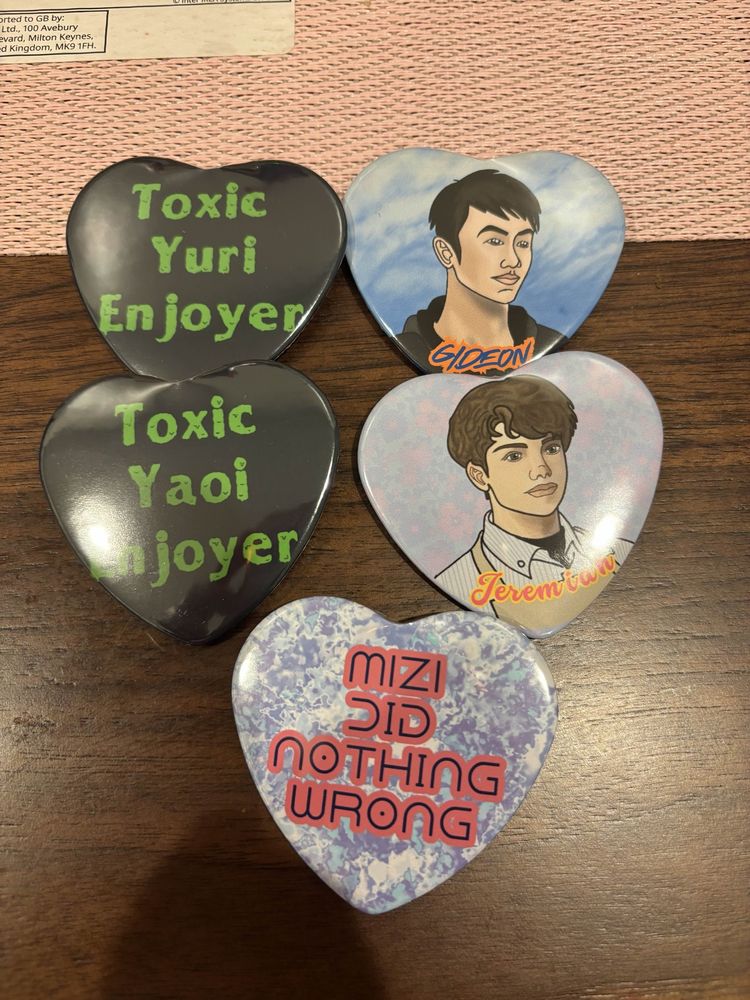 5 heart shaped button badges by corrin. Top left says toxic Yuri enjoyer, next to that is a drawing of Gideon from love and deepspace, middle left is toxic yaoi enjoyer, next to that is Jeremiah from love and deepspace, and the bottom is Mizi did nothing wrong (a reference to alien stage)
