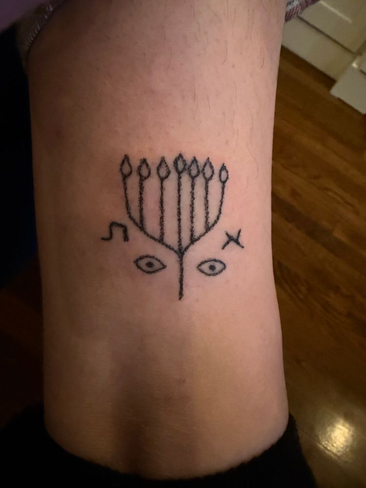A stick and poke tattoo of a design based on a menorah with two eyes best along the stem and aleph and tav on either side 
