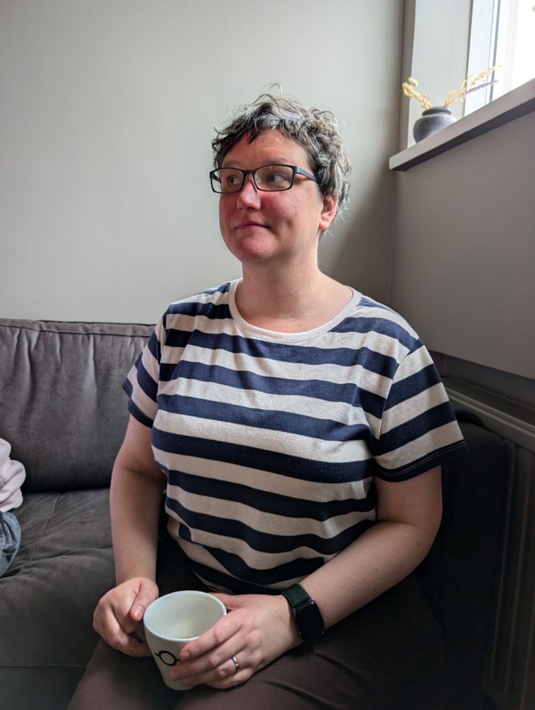 My frickin adorable wife in a blue and white striped shirt and black glasses, holding a coffee cup and looking off to her right with the cutest goddamn expression on her face. I want to squeeze this picture. 