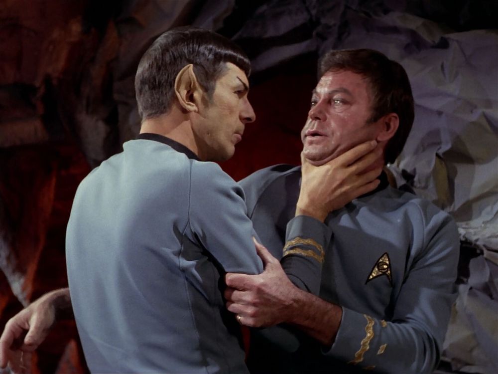 Spock and McCoy in a cave. Spock has seized McCoy by the throat angrily. 