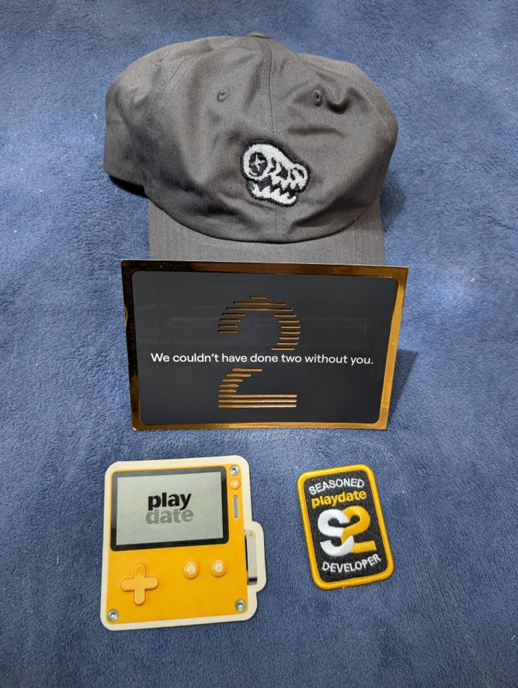 A picture of an official Dig Dig Dino cap (available in Playdate's Cotton Bureau), a sticker of the playdate, a thank you card from Panic for being a part of the second season and a patch that reads "seasoned playdate S2 developer".