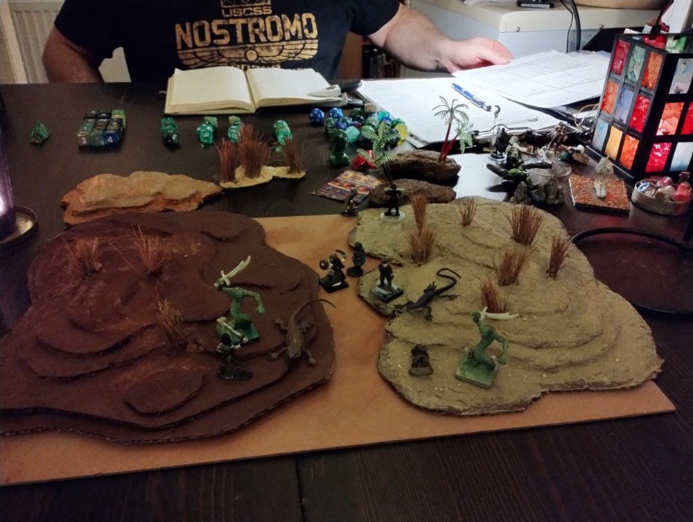 D&D terrain made to look like red clay desert dunes with patches of grass. Character minis and a variety of snakemen monsters are across the landscape in an epic battle scene. The background is a typical D&D session table crowded with dice, minis and candles.
