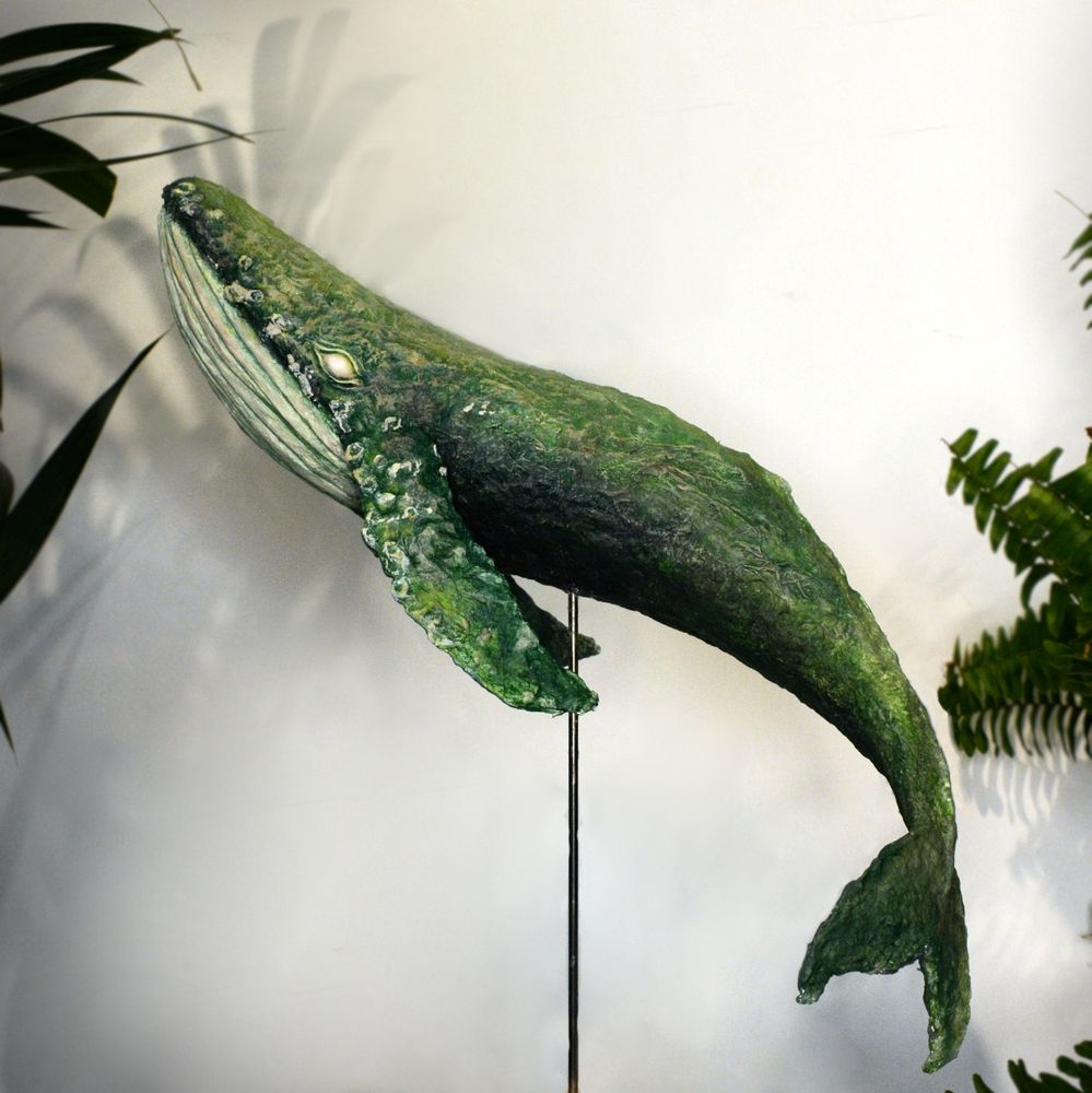 A paper mache humpback whale sculpture surrounded by houseplants against a white background. The whale is green and grey, similar to weathered stonework covered in moss.