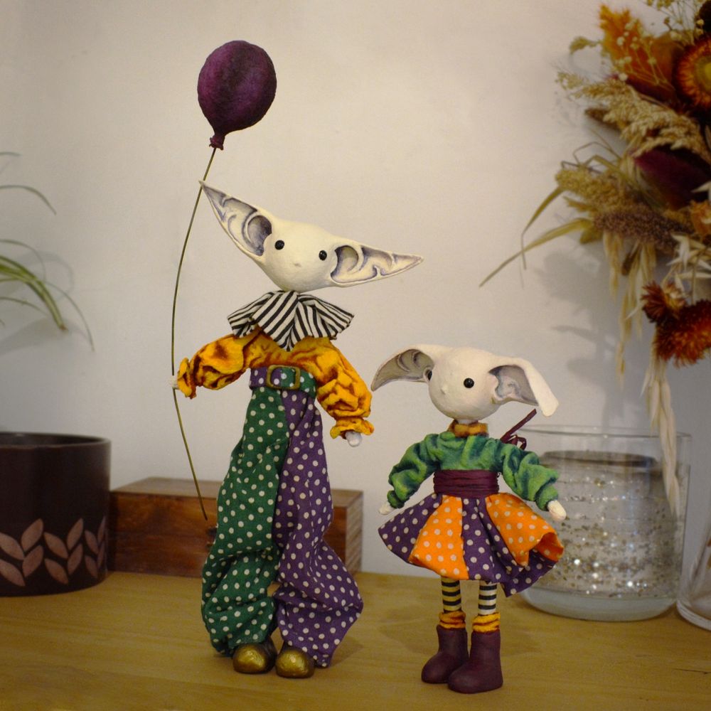 Two gremlin art dolls who might be siblings wearing bright, multicolored polka-dot clothes with circus vibes. They are made from paper mache, clay, and spun cotton, with clothes sewn from fabric scraps. One of them is taller than the other and holds a purple balloon; the smaller gremlin wears striped stockings and a circle skirt. They are standing on a wooden shelf in front of houseplants, dried flowers, and other decor.