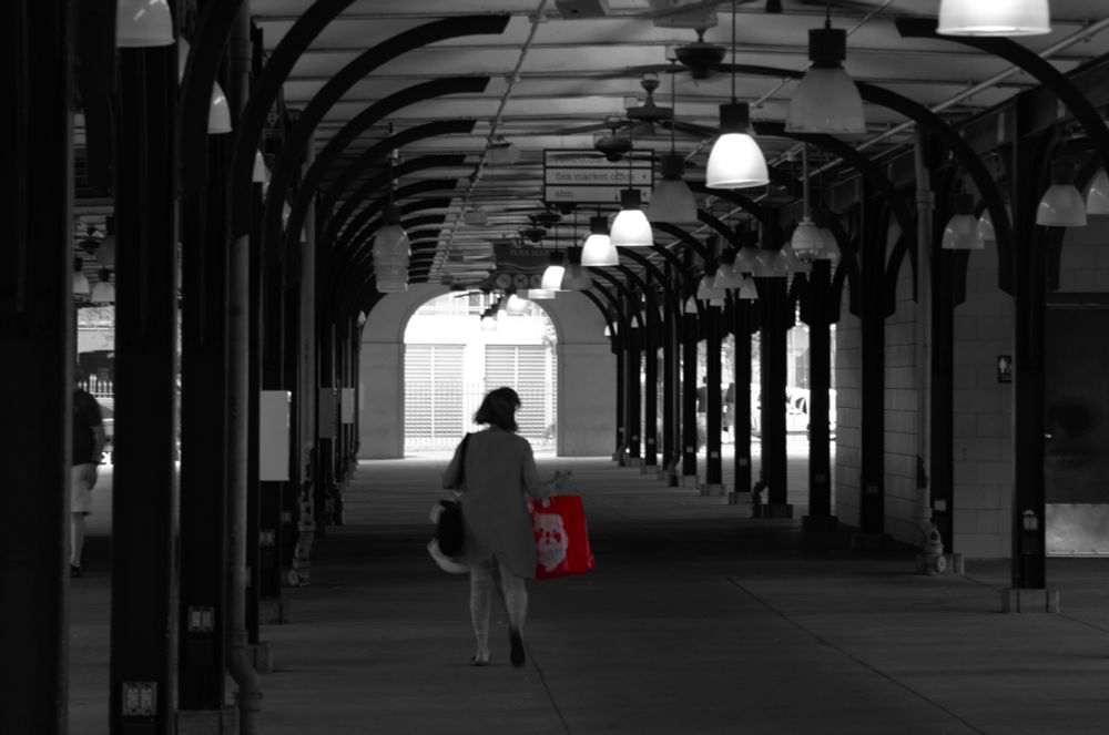 A person walks through the empty French Market building in the French Quarter, New Orleans. They're walking away from the camera carrying a bright red shopping bag. Everything else in the photo has been desaturated to black and white. The building is a roof held up by metal columns and has open carriage-style doorways.