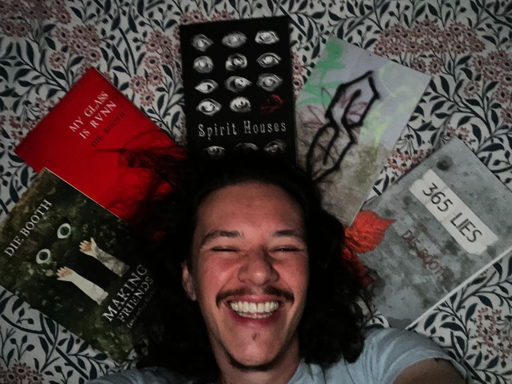 Author Die Booth, lying on a floral bedspread with his book titles (Making Friends, My Glass is Runn, Spirit Houses, Cool S and 365 Lies) spread out around his head. He is a middle aged white man with shoulder length dark hair and a dark moustache. He is grinning so widely that his eyes are closed.