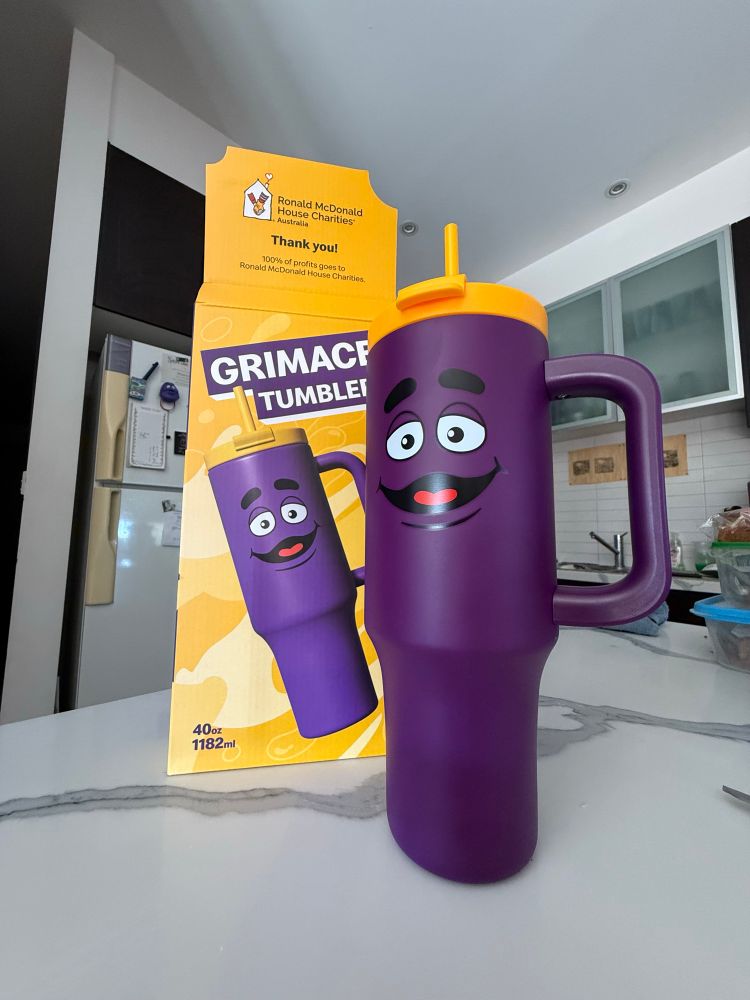 A purple tumbler with Grimace’s face on it from McDonalds. It has a yellow lid and a yellow straw. The cup sits in front of the box it came in. 