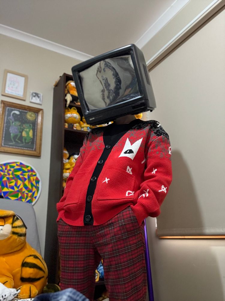 Tess showing off their new Cult of the Lamb cardigan. It is a delightful bright red with black trim and white accents. Tess is wearing their tv head and is hoping no one will mention it and pretend that this is completely normal.
