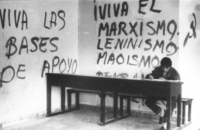 photo en noir et blanc d’un étudiant assis à une table de cours. tags sur le mur de gauche : « viva las bases de apoyo » (vive les bases d’aide). tags sur le mur du fond : viva el marxismo, leninismo, maoismo , [ illisible] » avec faucille et marteau
photo prise à l’université publique d’ayacucho durant le conflit armé péruvien (1980-2000)