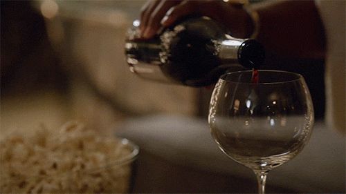 Imagen de alguien llenando una copa de vino. Se ve también un bol de palomitas encima de una mesa, delante de un sofá.