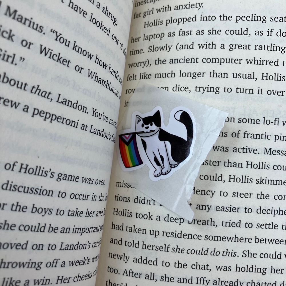 A photo of a book spread—pages 98-99of Christen Randall’s “The No-Girlfriend Rule.” In the crease is an unused sticker of a toony black and white cat holding a little pride flag in its mouth.