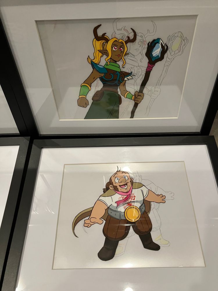A photo of two 11 x 14” matted frames, each with a mock anime cel inside, backed by a relevant douga/pencil drawing. This one features Darsi, a blonde wood elf with antlers and a staff, and Robertcat, a dwarf with a pink scarf and champion belt.