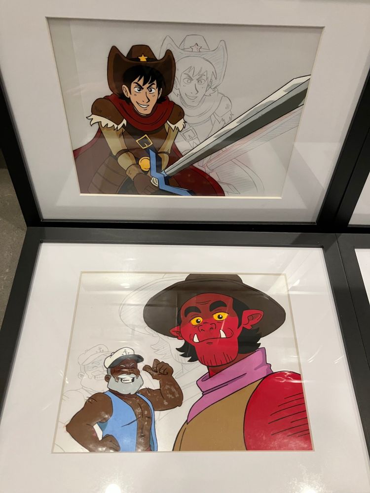 A photo of two 11 x 14” matted frames, each with a mock anime cel inside, backed by a relevant douga/pencil drawing. This one features Jonathan, a cowboy with a sword, and Skipper and Big Tex, a dark-skinned man with a sailors cap and a big red hobgoblin with a cowboy hat.