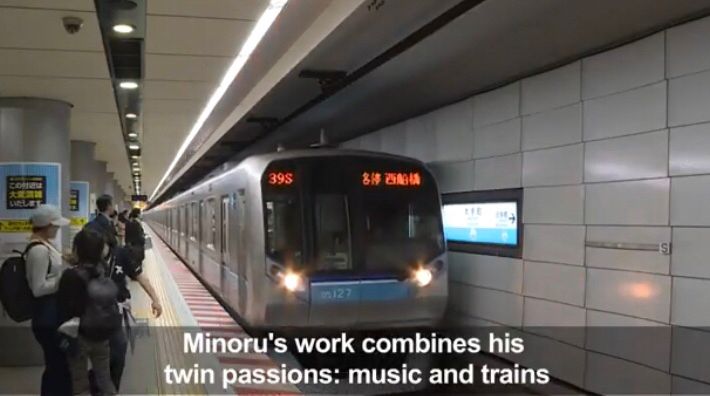 A sleek subway train is arriving at a modern underground station, labeled "39S" on its digital front display, alongside text in Japanese. The platform features clean, tiled walls and directional signage. Several passengers stand near the edge, including a woman wearing a white hat and backpack, awaiting boarding. Below the image, the caption reads: "Minoru's work combines his twin passions: music and trains."