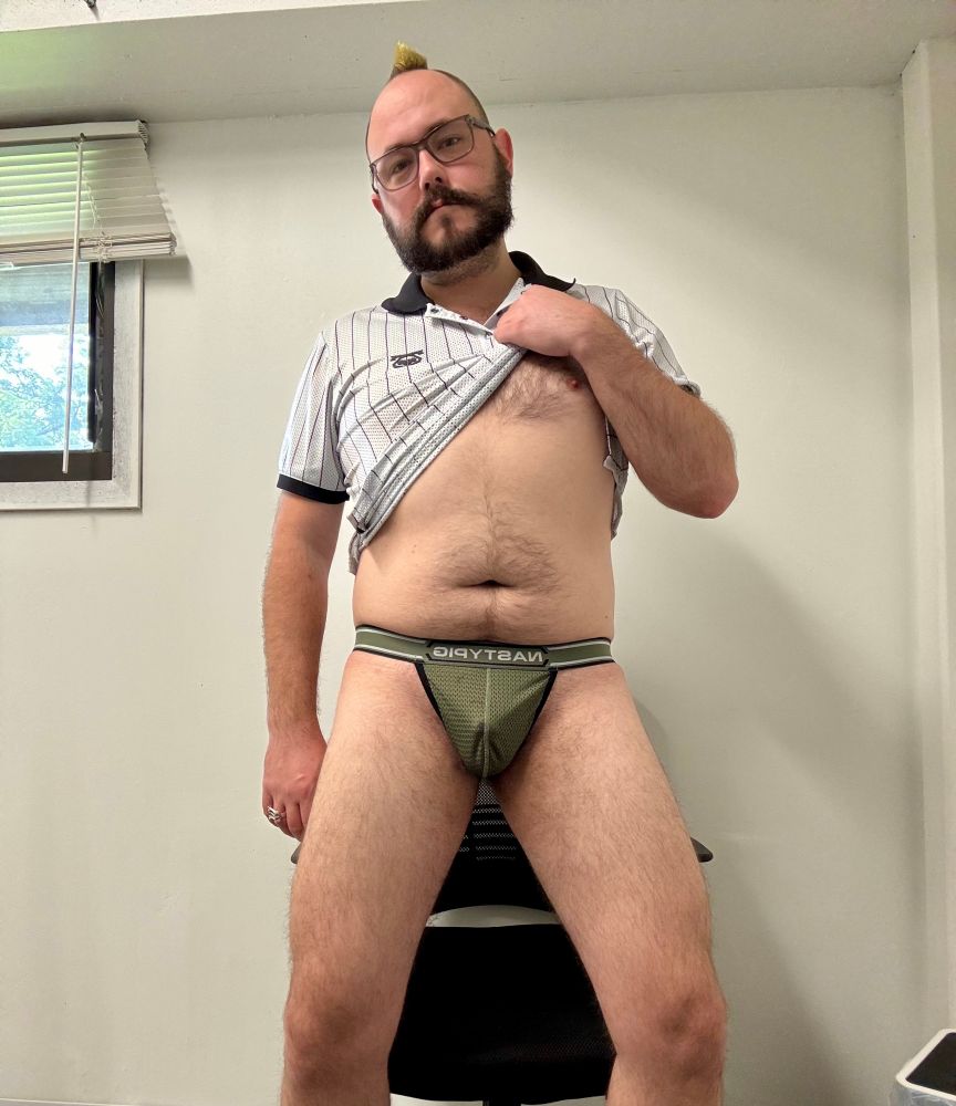 Faust wearing a military green mesh thong. He’s lifting up his mesh white and black striped polo and his pants are down. He’s in a plain office.