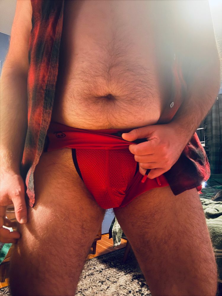 Faust wearing a red pair of athletic shorts with a mesh jockstrap attached underneath. His hand is holding the shorts to the side to reveal the jock.