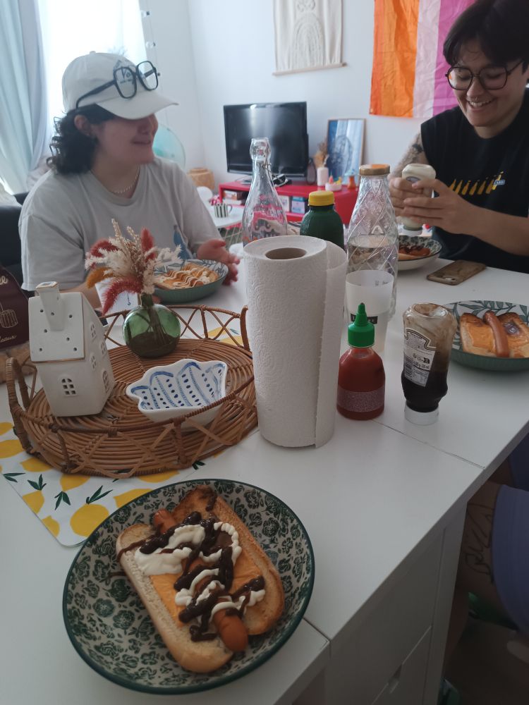 Table blanche en intérieur recouverte d'assiettes de hot-dogs, de sauces et de boissons. Jo en face à gauche et lena et face à droite qui sourient. Bout de la jambe de Noé assis à ma droite