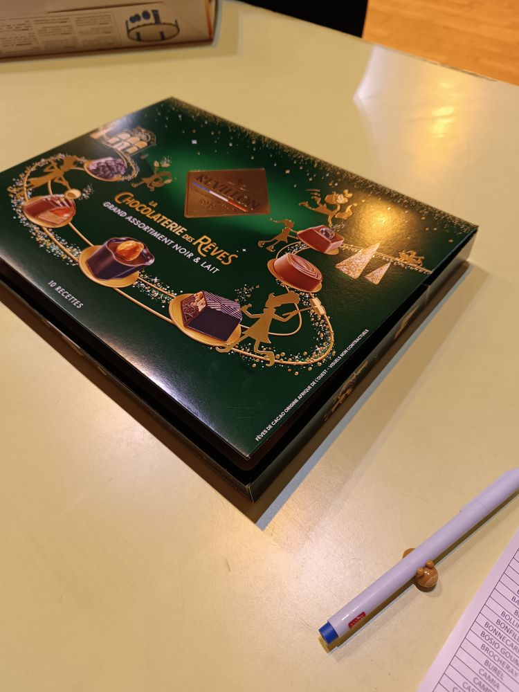 Boite de chocolats posée sur une table jaune en intérieur à côté d'un stylo legamy paresseux et d'une liste d'appel