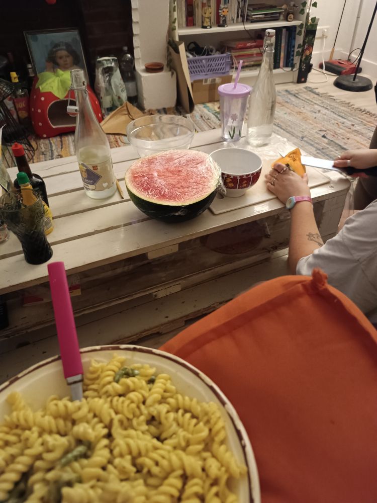Assiette de pâtes sur mes genoux dans le canapé, jo à ma droite assis.e par terre en train de découper de la pastèque et du melon 
