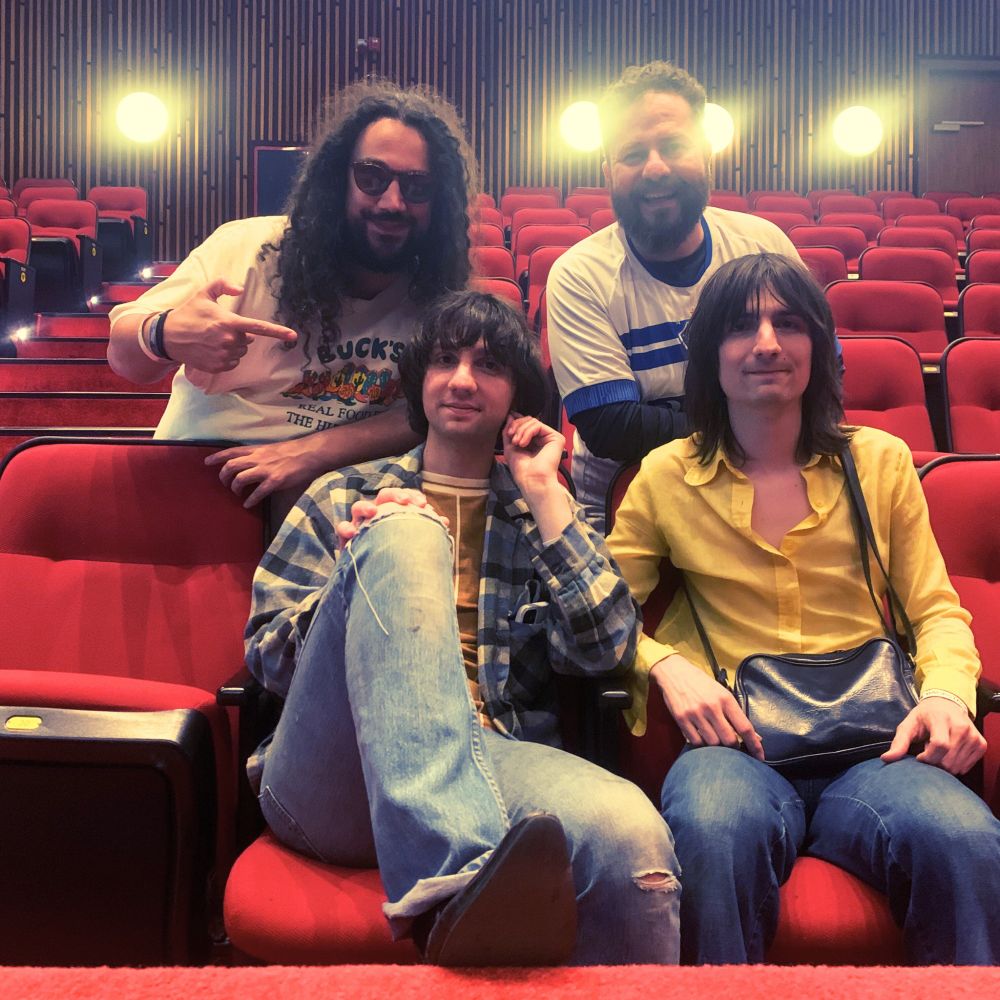 Quatro homens sentados nas cadeiras de um teatro com poltronas vermelhas. Os dois de cima são Bruno Capelas (homem de cabelos cacheados, com óculos escuros, barba e camiseta branca) e Igor Muller (ruivo, com barba ruiva e uma camiseta azul e branca do time do Paysandu). Os dois sorriem. Bruno aponta para os homens da fileira de baixo: Michael Daddario e Brian Daddario, os dois irmãos que fazem parte da banda Lemon Twigs. Michael usa uma camisa xadrez azul, uma calça jeans e uma camiseta marrom. Ele tem cabelosna altura do pescoço. Já Brian tem cabelos compridos, na altura dos ombros, usa uma camisa amarela, calça jeans e uma bolsa transpassada no colo. Michael tem um de seus pés apoiados na barreira do teatro. Ao fundo, refletores estão acesos. 