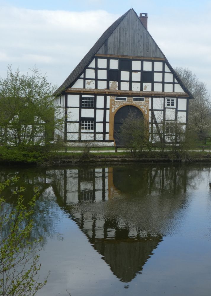 Fachwerkhaus, mit schwarzen Balken und weiß verputzem Fachwerk, spiegelt sich in einem davorliegenden See.