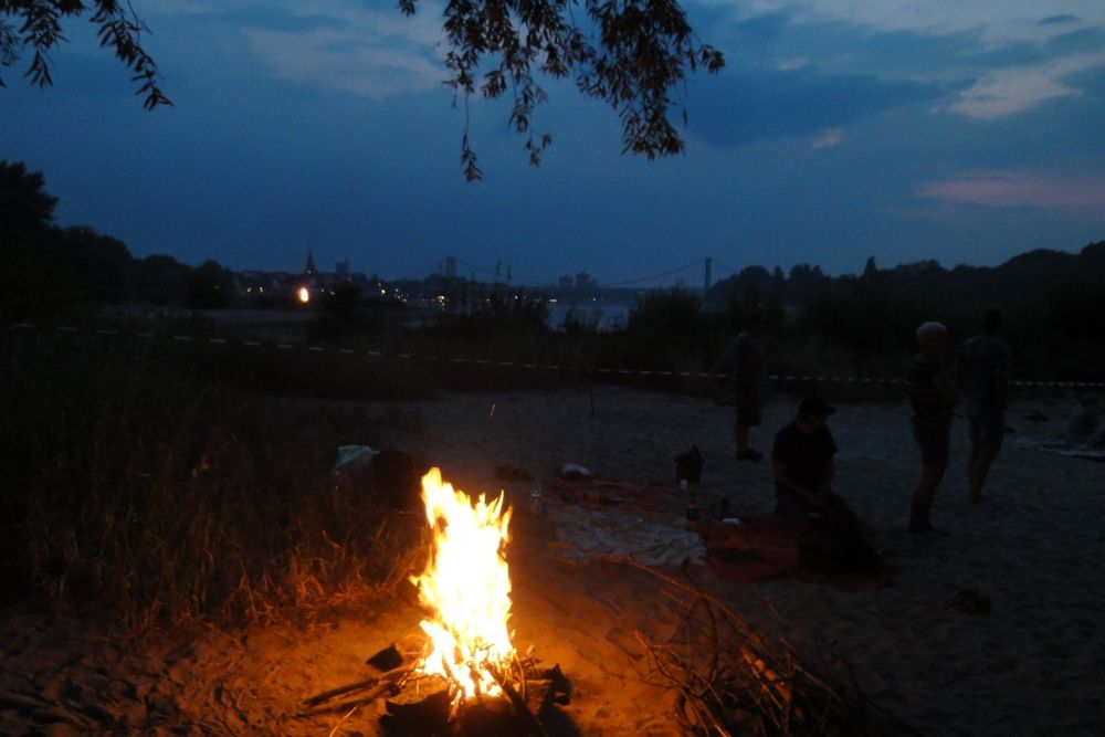 Am unteren Bildrsnd ein Lagerfeuer am sandigen Ufer eines Flusses. In der Nähe Menschen, als schwarze Schemen erkennbar.
In der Mitte der Fluss, dahinter Häuser, alles sehr dunkel.
Darüber graublauer, bewölkt Himmel.