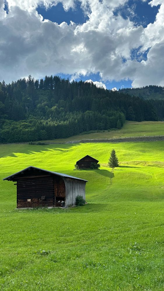 An einem grünen Berghang stehen zwei Berghütten in unterschiedlichen Höhen. Hin Hintergrund ist Wald zu sehen, der Himmel ist heiter bis wolkig. Durch die Wolken scheint die Sonne und hinterlässt helle Flecken auf dem grünen Hang.