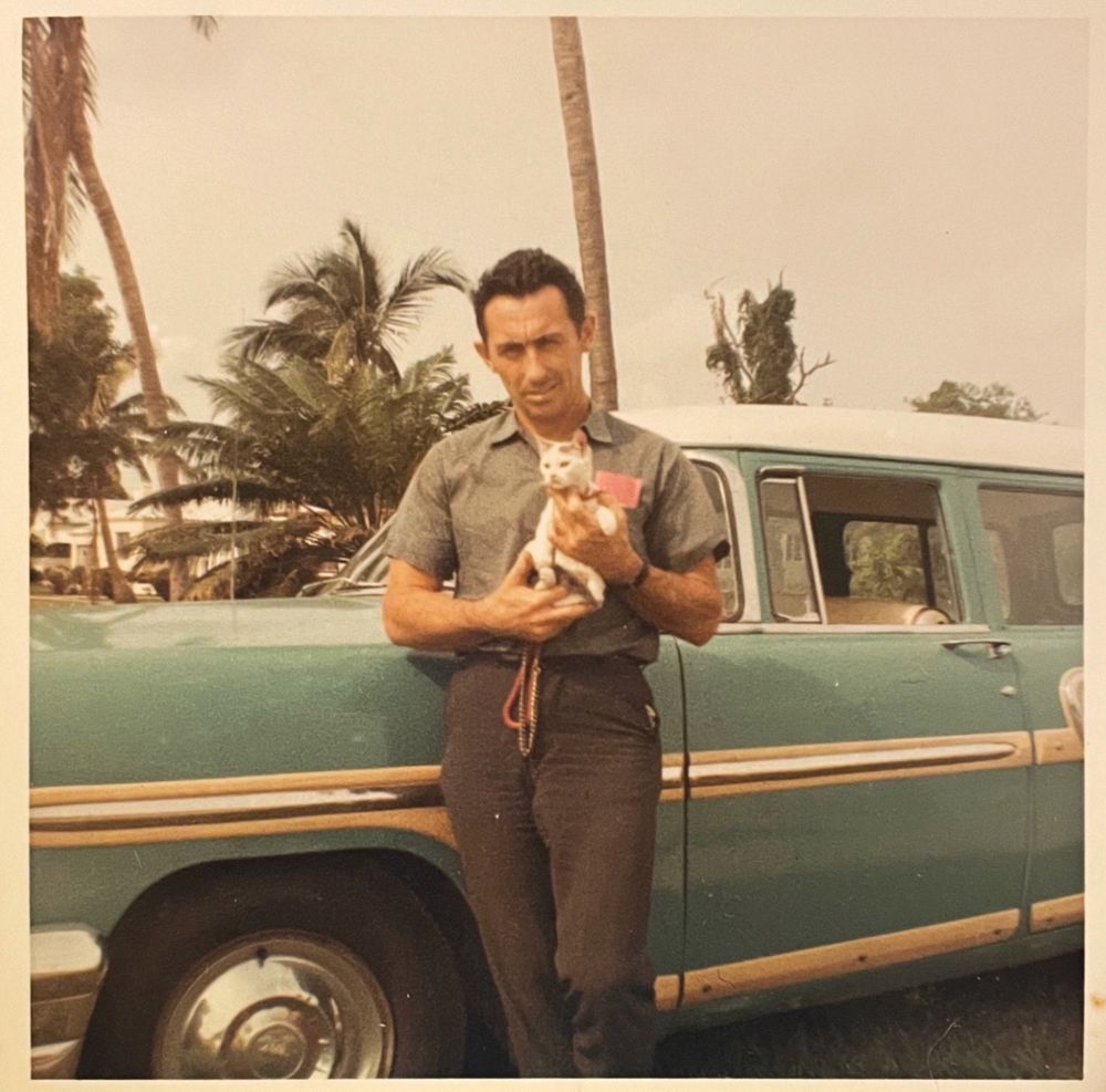 Color photo of a man with brown hair and tan or brown skin, leaning against a very cool huge teal car. In his hands he’s holding a tiny white and ginger kitten that seems like it might be wearing a collar and leash but it’s difficult to tell for sure. Palm trees rise up in the background.