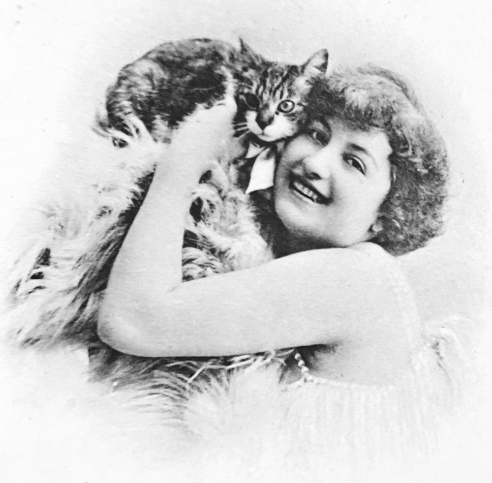Black and white photo of a light skinned woman smiling and adoringly holding a shorthaired tabby up, pressing it against the side of her face. The cat looks mildly terrorized.