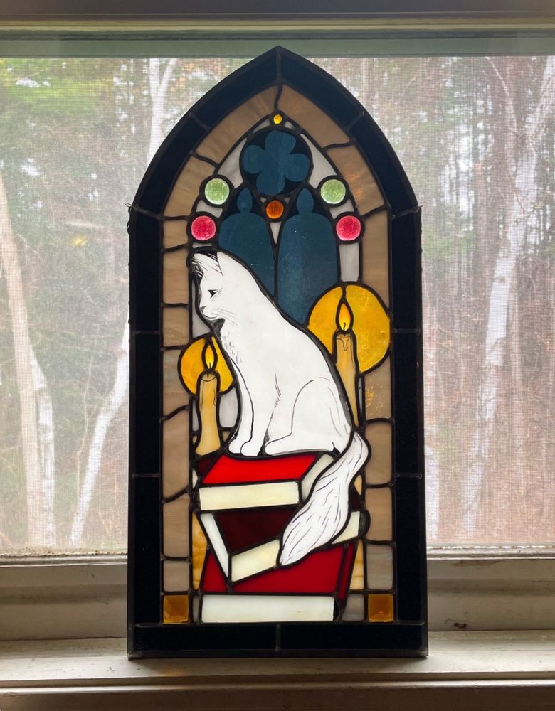 A stained glass piece about 2 ft tall and 1 ft wide, depicting a medium haired white cat sitting on top of a stack of large hardcover books, two off white candles glowing nearby. In the background you see tall, arched church windows with a round window above them. The glass set in the windows is a mellow dark blue like the night sky. That in combination with the candles gives the scene a feeling of cozy nighttime.