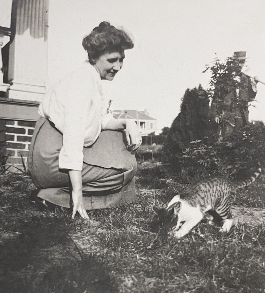 Black and white photo of a smiling white woman in Edwardian clothing, kneeling on a grass lawn to admire a white and tabby shorthaired cat. A man approaches from the background on the right.