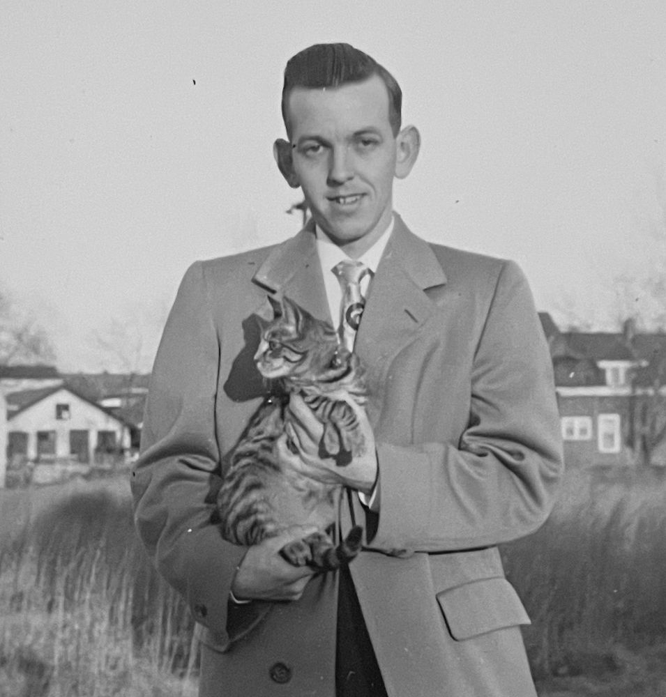 Black-and-white photo of a young light skinned man standing outside wearing an overcoat and he seems to be wearing a suit underneath that. He’s smiling at the camera and holding up a shorthaired tabby cat as if showing it off.