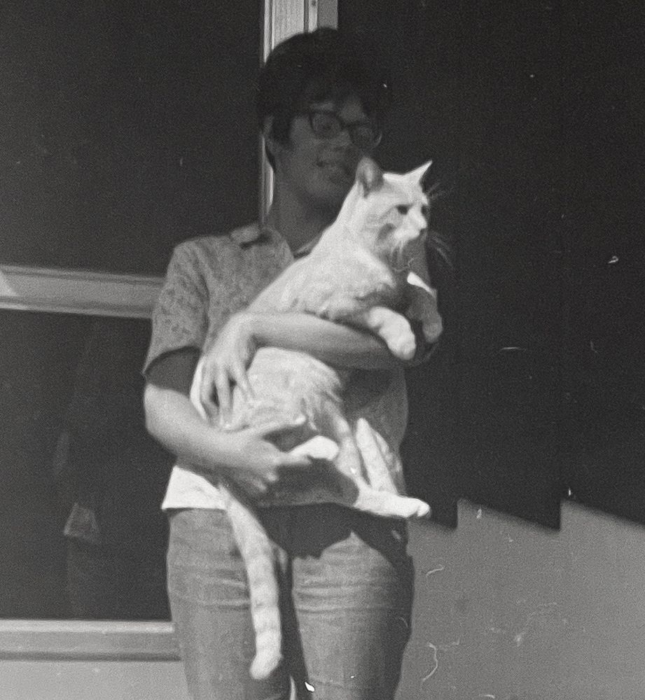 Black and white photo of a white woman standing outside of a house, holding a shorthaired ginger tabby. The woman’s face is in shadow but the cat is held in the sunlight.