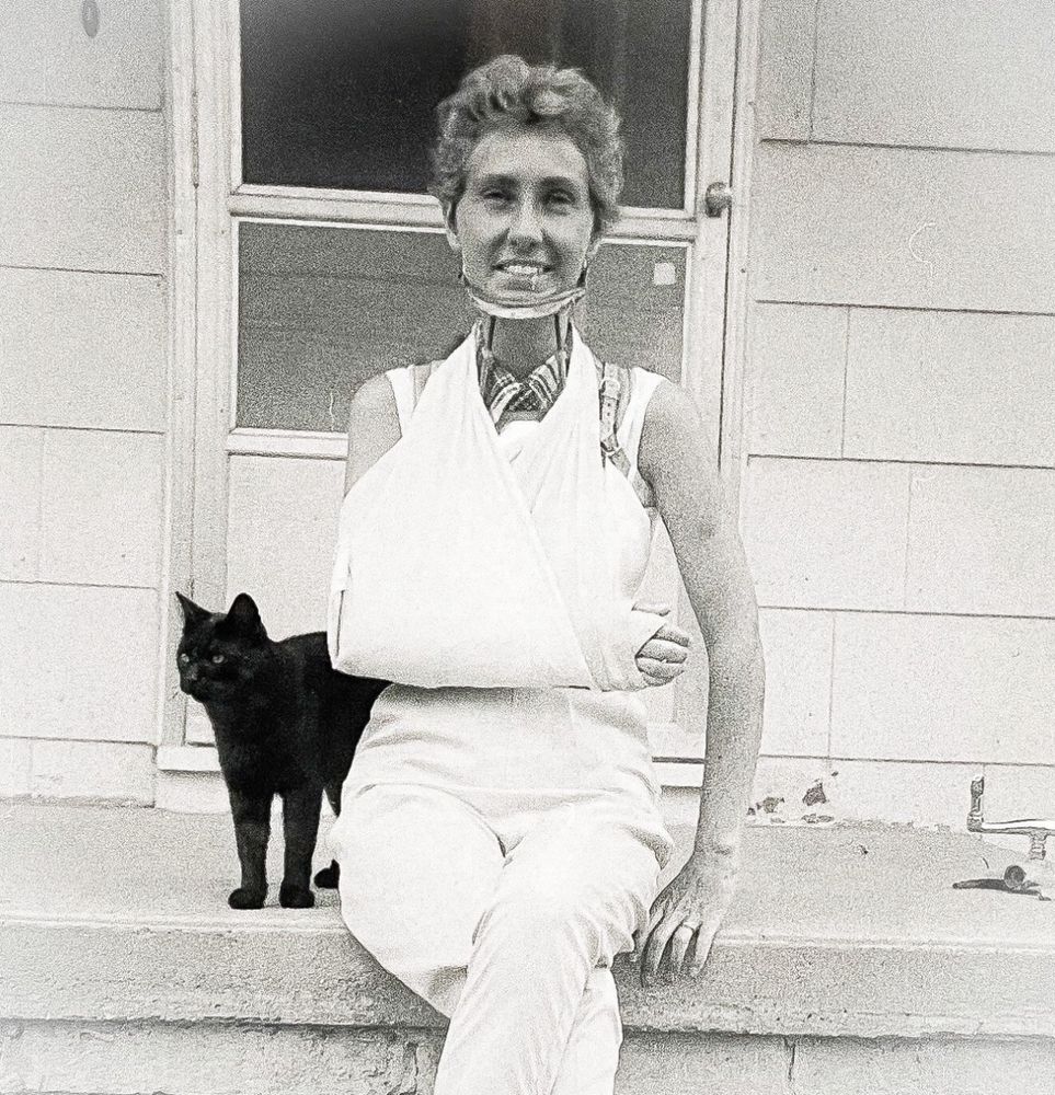 Black and white photo of a smiling white woman sitting outside on a sunny house stoop, wearing a cast and sling on her right arm and cervical collar. A happy little shorthaired black cat rubs up beside her.