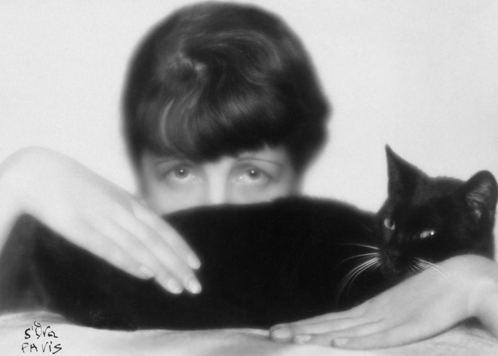 A white woman with dark hair peeks over a glossy black cat that is lying on a table in front of her. You can only see the woman's eyes and she gazes intensely at the camera as her hands stroke the beautiful, relaxed cat.