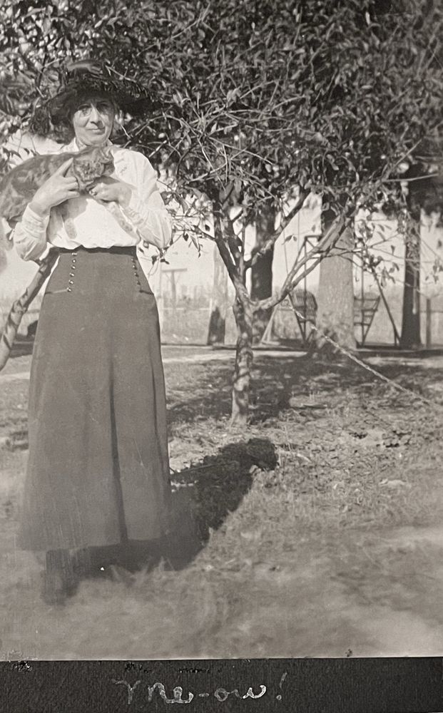 Black-and-white photo of a light skinned woman wearing a dark hat, white blouse and a long dark skirt. Her clothing style looks to be roughly Edwardian. She is standing outside holding a short haired tabby cat that looks like it is probably ginger. The photo is mounted to black paper and written at the bottom in cursive writing is the word meow with an exclamation point.