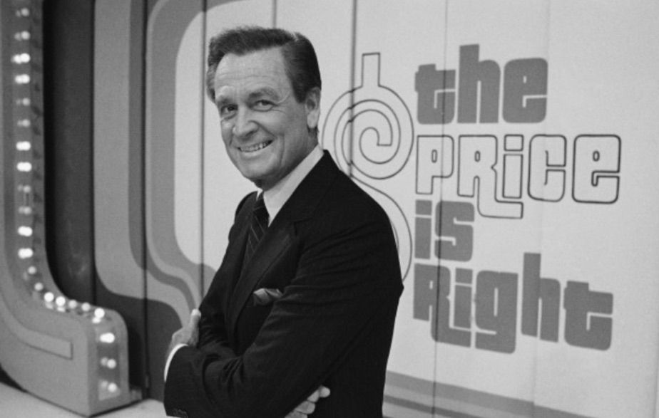 Black and white photo of game show host Bob Barker, a middle aged white man wearing a dark suit. He is standing on the set of The Price Is Right, its logo prominently displayed on the wall behind him.