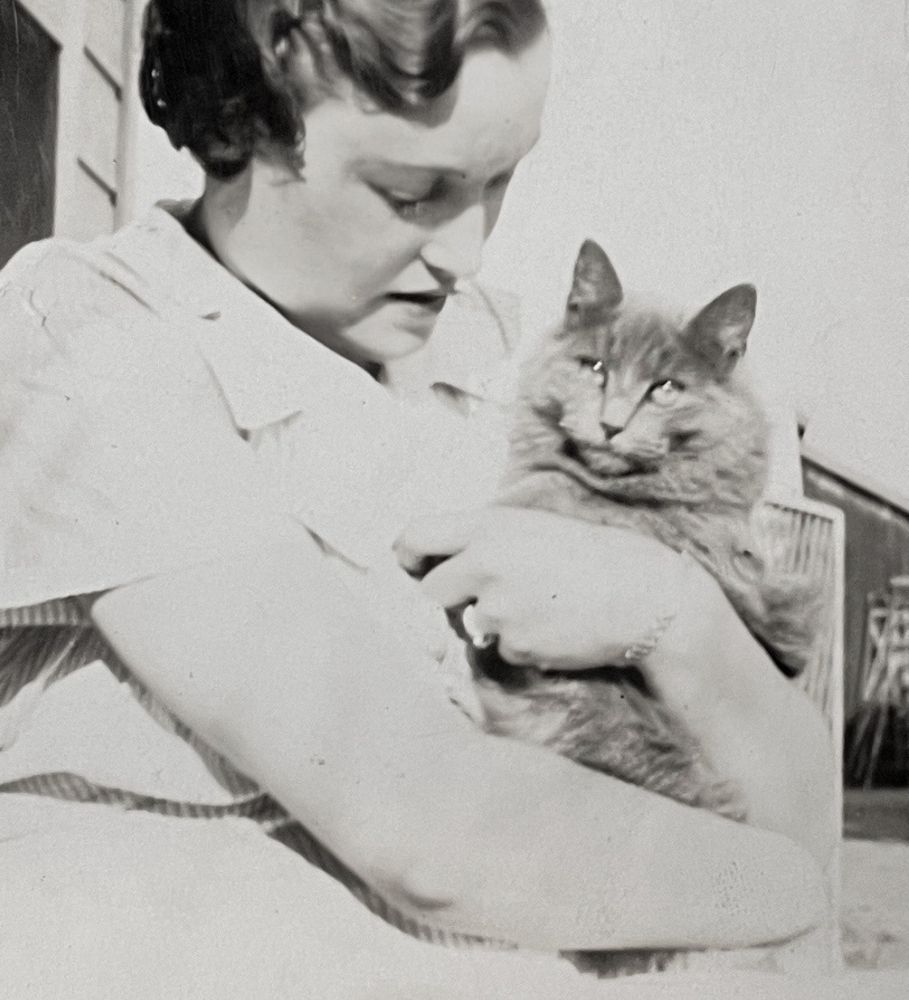 Black and white photo of a white woman with dark wavy hair, looking down affectionately at a longhaired grey cat that she cradles in her arms like a baby.