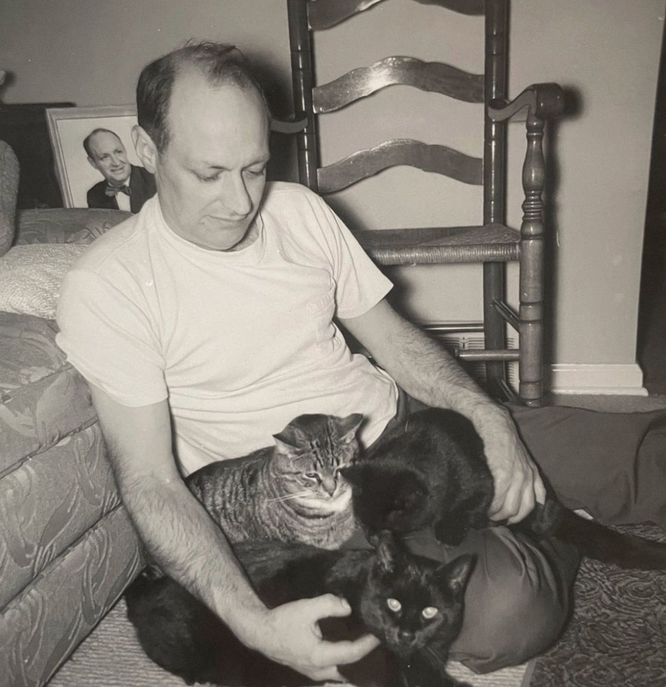 Black and white photo of a balding white man sitting on the floor in front of a couch. He is looking down at three cats. A shorthaired tabby and shorthaired black cat are on his lap and he’s holding another black one against the side of his leg. There seems to be a framed photo of the man wearing a bowtie on a side table in the background.